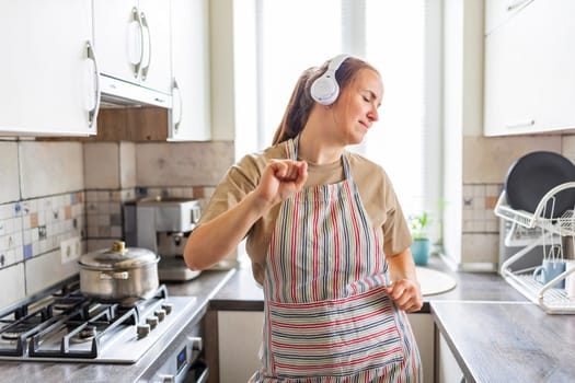 Smiling beautiful woman dancing in kitchen listening music with headphones.