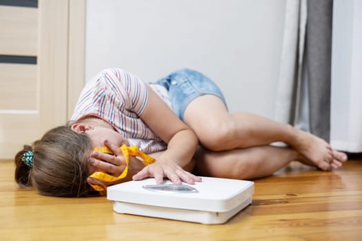 Scales and depressed young woman lying on the floor holding her head and covering her face with her hands.