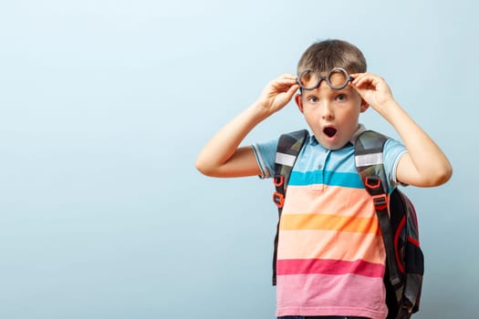 Surprised schoolboy in glasses with backpack on blue background. Back to school. Mockup, copy space.