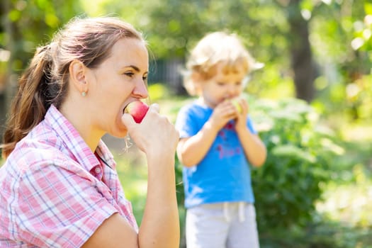 Mother and her cute little son eating ripe red apples in beautiful garden. Healthy food concept.