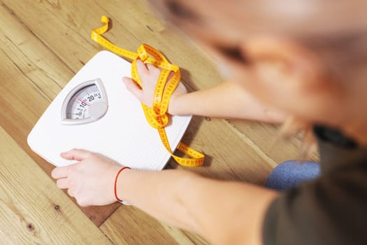 Depressed, frustrated and sad woman with a measuring tape looks at a white scale on a wooden floor.