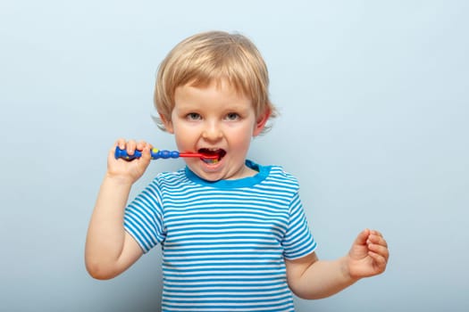 Little blonde boy brushing teeth with toothbrush on blue background. Dental hygiene.