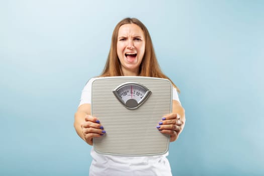 Frustrated young brunette woman in a white t-shirt with weight scale on blue background.