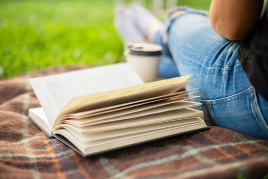 A woman in jeans with a book and a cup of coffee sits on a blanket in the park.
