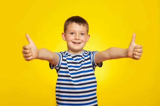 Portrait of happy boy in striped t-shirt showing thumbs up gesture on yellow background.