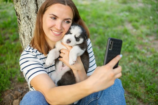 Young beautiful woman in a t-shirt and jeans with a husky puppy takes a photo or selfie on the phone while sitting in a park near a tree.