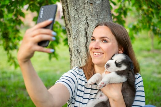 Young beautiful woman in a t-shirt and jeans with a husky puppy takes a photo or selfie on the phone while sitting in a park near a tree.