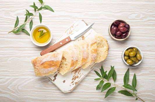 Sliced fresh ciabatta on cutting kitchen board, green and brown olives, olive oil with rosemary, olive tree branches, white wooden rustic background top view