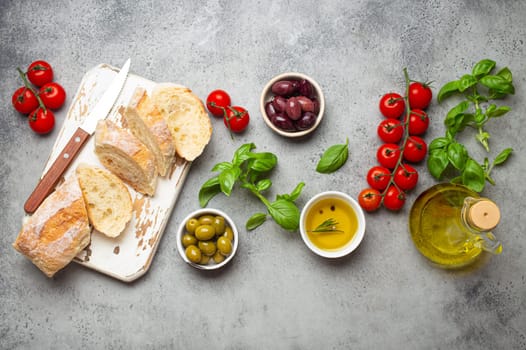 Food composition with sliced ciabatta, olives, olive oil, spaghetti, fresh basil, cherry tomatoes on gray concrete stone rustic background top view. Italian cuisine concept