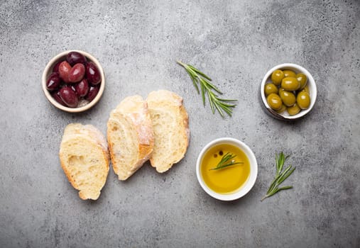 Slices of fresh ciabatta, green and brown olives, olive oil with rosemary, olive tree branches on gray concrete stone rustic background overhead