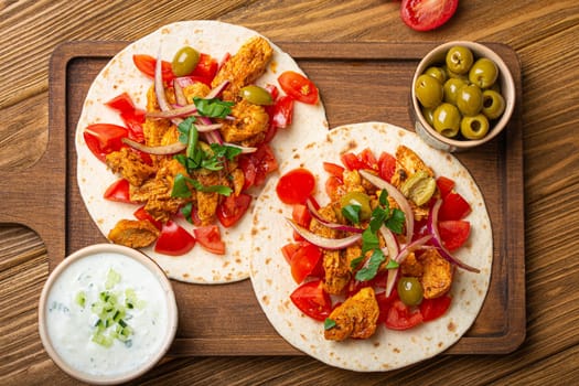 Cooking Traditional Greek Dish Gyros: Pita bread with vegetables, meat, herbs, olives on rustic wooden cutting board with Tzatziki sauce, olive oil top view, wooden light brown background.