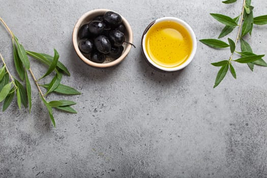 Black olives and extra virgin olive oil in little bowl with olive tree branches top view on gray concrete stone background, space for text