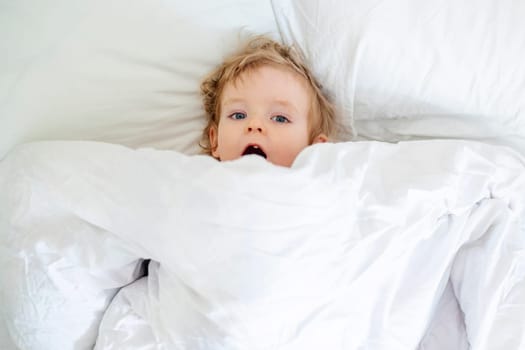 Top view of cheerful little boy in white bed early in the morning. Portrait of a handsome boy awakened.