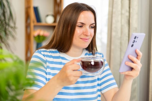 Happy beautiful woman sitting at table with cup of tea and chatting with friends on social networks, playing game or buying online