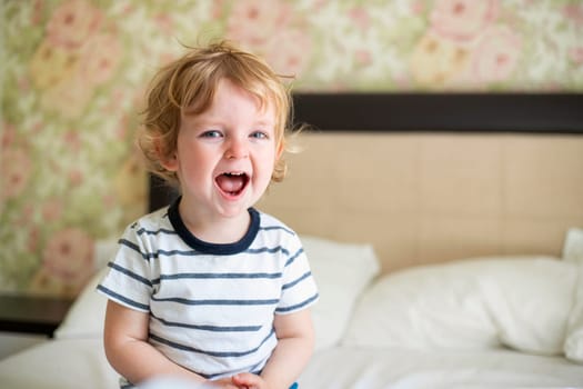 Cute laughing child boy sits in bed. Boy plays at home. Child in a sunny bedroom.