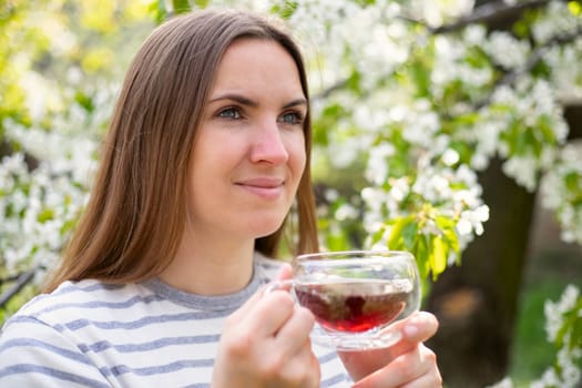 Young woman drinking tea in glass cup among the flowering trees in the garden.