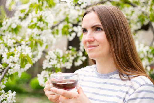 Young woman drinking tea in glass cup among the flowering trees in the garden.