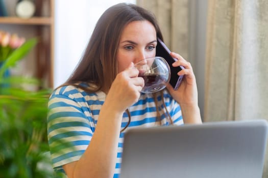 Connection and communication. Brunette female working from home using laptop speaking on mobile phone and drinking tea. Distance freelance job concept