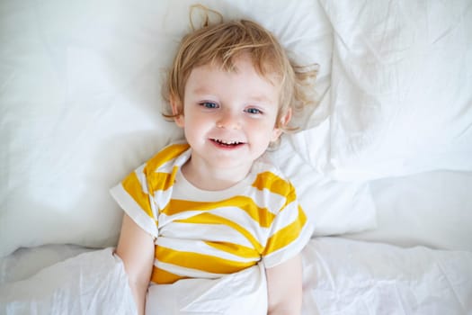 Top view of cheerful little boy in white bed early in the morning. Portrait of a handsome boy awakened.