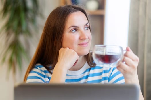 Smiling woman taking a tea break as she sits at a table working on a laptop computer.