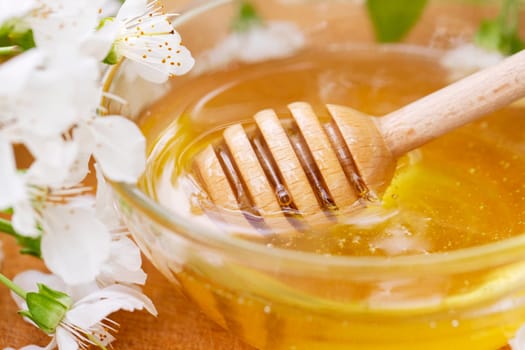 Flower honey in a glass bowl with wooden honey spoon on wooden table with blossoming flowers.