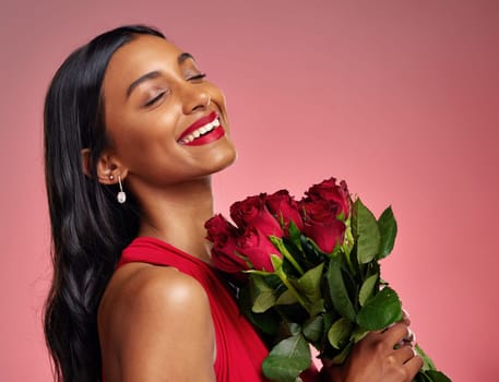 Face, beauty and smile of a woman with a roses on a studio background for valentines day. Makeup, model and happy young Indian girl with a flower bouquet for romance or love on a pink backdrop.