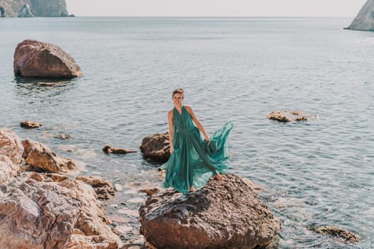 Woman green dress sea. Woman in a long mint dress posing on a beach with rocks on sunny day. Girl on the nature on blue sky background