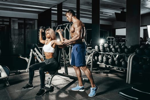 Fitness instructor exercising with his client at the gym. The beautiful blonde is engaged in sports. Handsome man, sports instructor