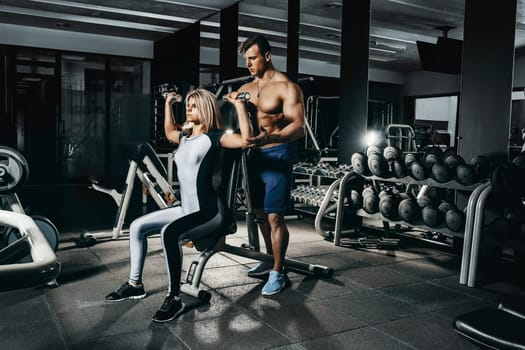 Fitness instructor exercising with his client at the gym. The beautiful blonde is engaged in sports. Handsome man, sports instructor