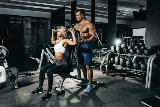 Fitness instructor exercising with his client at the gym. The beautiful blonde is engaged in sports. Handsome man, sports instructor