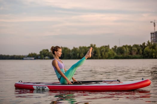 Young woman sitting on paddle board, practicing yoga pose. Doing yoga exercise on sup board, active summer rest. Exercise for flexibility and stretching of muscles.