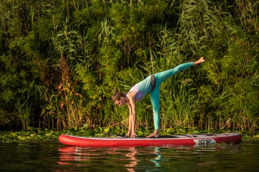 Photo of young woman doing hand stand on stand up paddle board. She wearing a leggings and top. Sunny day, blue lake and green trees on the shore.