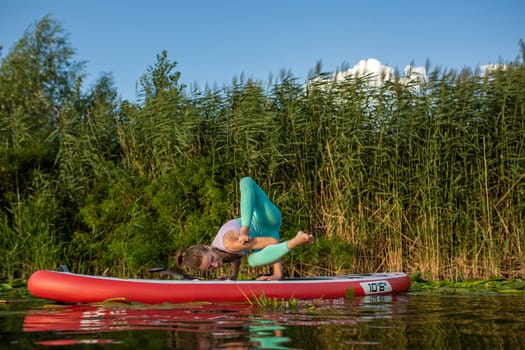 Photo of young woman doing hand stand on stand up paddle board. She wearing a leggings and top. Sunny day, blue lake and green trees on the shore.