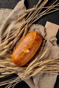 Whole loaf of freshly baked white bread on cut of burlap with ears of wheat and sheet of craft paper with empty space for text, top view on black background. Artisanal breadmaking concept