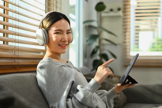 Cheerful asian woman wearing wireless headphone and enjoying browsing internet on digital tablet at home.