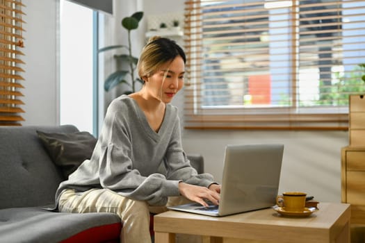 Pleasant asian woman wearing casual clothes sitting on couch, working remotely, surfing internet on laptop at home.