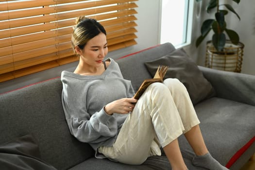 Carefree asian woman sitting on comfortable couch and reading book. People, leisure and lifestyle concept.