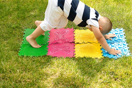 A little boy goes in for sports on massage orthopedic multi-colored rugs. A child in a striped T-shirt crawls merrily in the fresh air in the park, on green grass on massage mats. Stands in a plank position.