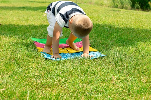 A little boy goes in for sports on massage orthopedic multi-colored rugs. A child in a striped T-shirt crawls merrily in the fresh air in the park, on green grass on massage mats. Stands in a plank position.