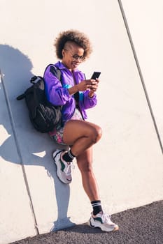 Vertical portrait of a stylish african american sportswoman using a mobile phone outdoors, concept of technology of communication and urban sports
