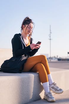 vertical photo of a sportive woman sitting outdoors in the city using a mobile phone, concept of technology of communication and urban lifestyle, copy space for text