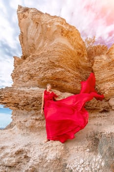 woman red silk dress stands by the ocean, with mountains in the background, as her dress sways in the breeze