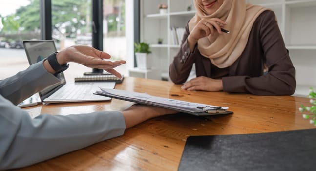 Young muslim female business worker discussing with friends sitting together, working with laptop and financial documents at desk.
