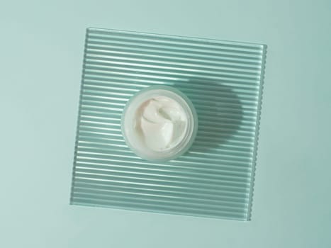 Cosmetic cream or moustirizer on transparent ribbed acrylic plate over blue background. Open round glass jar with aesthetic swirls cream. Top view flatlay. Copy space
