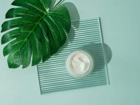 Cosmetic cream or moustirizer on transparent ribbed acrylic plate with tropical palm monstera leaf over blue background. Open round jar, aesthetic swirls cream. Top view flatlay. Copy space