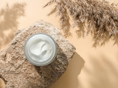 Cosmetic cream or moustirizer on stone, pampas grass on pastel beige background. Beautiful shadows. Open round glass jar with aesthetic swirls face cream. Top view or flat lay. Copy space