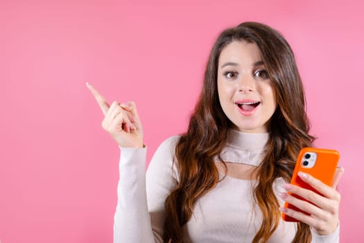With a smile, a lovely woman captures attention as she showcases the copy space, holding a mobile phone against pink backdrop. Advertising concept.