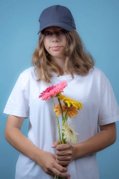 portrait of a pretty Caucasian girl with gerbera flowers on a blue background. A teenage girl in a white T-shirt and baseball cap holds flowers and looks at the camera