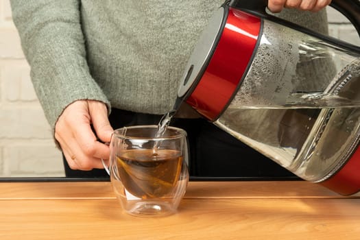 Woman poring hot boiled water from electric kettle into a clear glass cup on table. The process of brewing tea or tea ceremony in a warm soft light. Movement of boiling water pouring into a cup