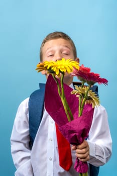 a first grader in a white shirt with a backpack and closed eyes holds a bouquet of flowers in his hands. Cute Caucasian boy goes to school with a bouquet of flowers and a backpack. Side view. Schoolboy. September 1, for the first time in first grade and, blue background, vertical photo